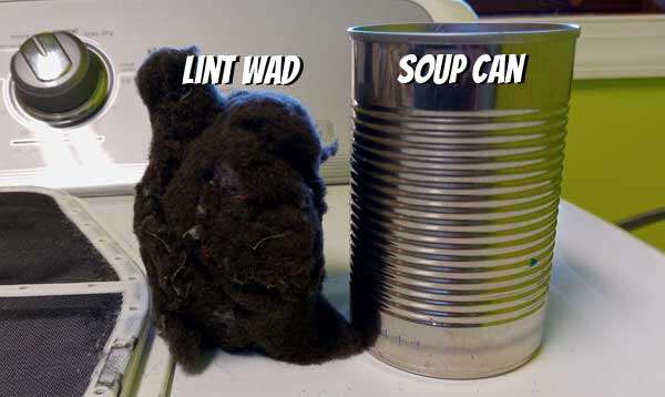 A wad of dark lint standing next to a soup can atop a clothes dryer. They are approximately the same size.