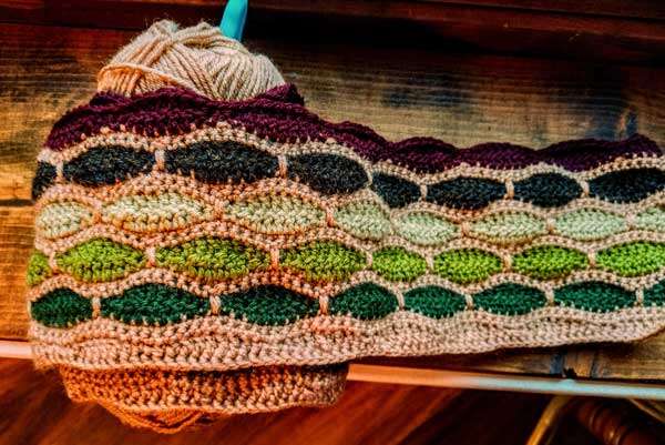Strip of crochet with rows of slightly rounded bricks in three shades of green, dark brown, and burgundy, separated by beige grout.