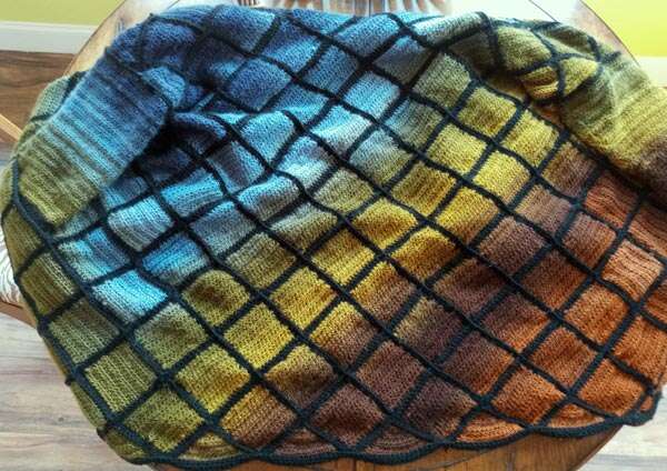 Patchwork crocheted sweater spread out on a table. Small squares are arranged on the bias, like diamonds, with a foresty color gradient blending dark blue, light blue, mossy green, golden green, chocolate brown, and copper. Black seams separate each square.