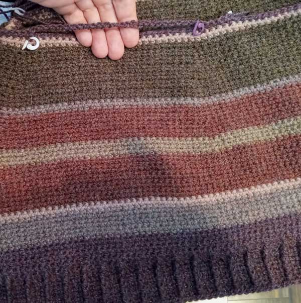 Crochet project in progress with stripes of purple, magenta, and brown. A white woman's fingers poke through a gap that will become a pocket.