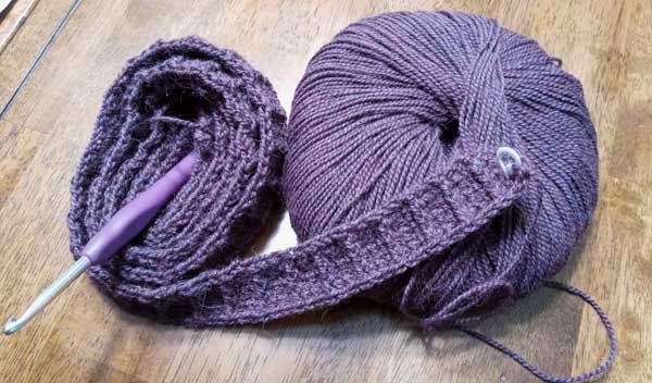 A ball of purple yarn beside a coil of crochet trim about an inch tall with raised vertical ribbing.