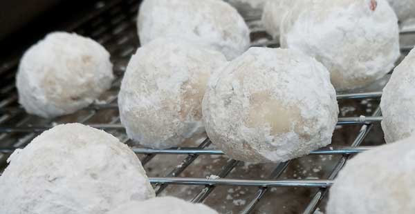 Small orbs of pecan shortbread rolled in powdered sugar resting on a wire rack. The sugar fallout below the rack foretells the state of one's shirt after eating these.