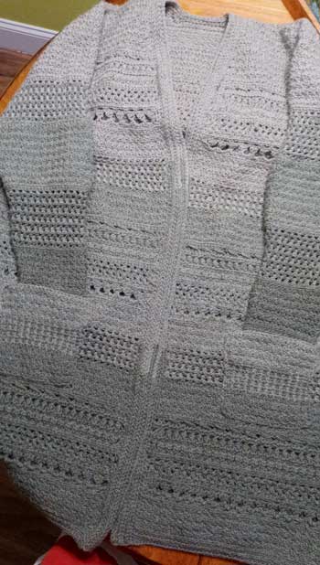 A long cardigan with three blocks of color, lightest at the shoulders, darkest at the bottom. Various types of stitches create a striped effect. The sleeves are tucked into pockets.